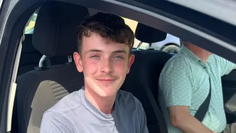 BBC/Emily Johnson A teenage boy with short dark hair smiles from the driver's seat of a car.
