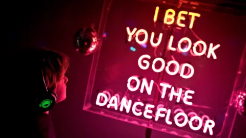 A pink lit up sign which reads 'I bet you look good on the dancefloor' in a clear, square casing. There is a small disco ball to the left and a young boy with short hair and lit up over-the-ear headphones looking at the sign. He is partly in shadow. The photo is slightly on an angle and the sign is lighting up the room in a pink light.
