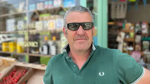 George Carden/BBC Eamon Stack standing outside his shop wearing a green polo top and sunglasses. Behind him is fresh produce displayed outside the shop