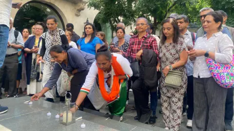 Photo of two women leaning down to place votives on some stone paving outside the embassy. There are people standing behind them and the embassy building in the background.