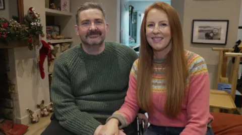 Dan and Anna smiling in their living room. Dan (left) is wearing a green jumper and Anna (right) is in a pink winter jumper with yellow and blue pattern.