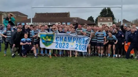 Kingwood RFC men in their sky blue and dark brown hooped rugby shirts. They are hold a banner proclaiming them 2023/2024 Gloucestershire Tribute South champions. All are smiling and several are punching the air in celebration.