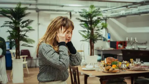 Chuko Cribb Nia Roberts has her head in her hands and has a pained expression on her face. She is stood in what looks like a kitchen with food and kitchen utensils seen on her table to the right. There are two tress in the background as part of the set.