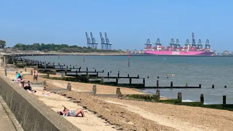 BBC Weather Watchers/duke skywalk An expanse of yellow sand at Harwich, with rows of wooden groynes running into the blue sea. A woman is lying on her back in the middle ground, near the sea wall, and there are a few other people on the beach. In the distance is a bright pink container ship. 