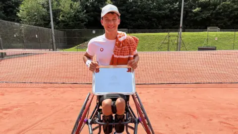 Leanne Rinne/BBC A teenage boy in a wheelchair holding a silver trophy shield on a tennis court 
