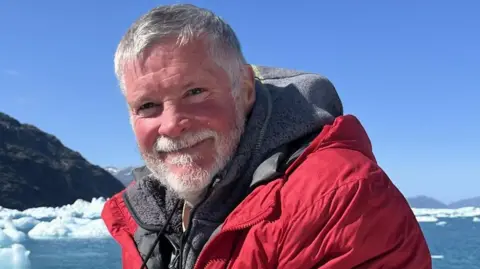 John O'Raw John O'Raw has short grey hair and a beard. He is wearing a large grey hoodie and red overcoat. He is smiling towards the camera and behind him is blue sea with ice flows and mountains in the distance. 