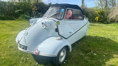 Wynford Jones A man sits in a small car, which can only fit a single occupant in, with a bubble-style body. It's parked on grass on a sunny day.