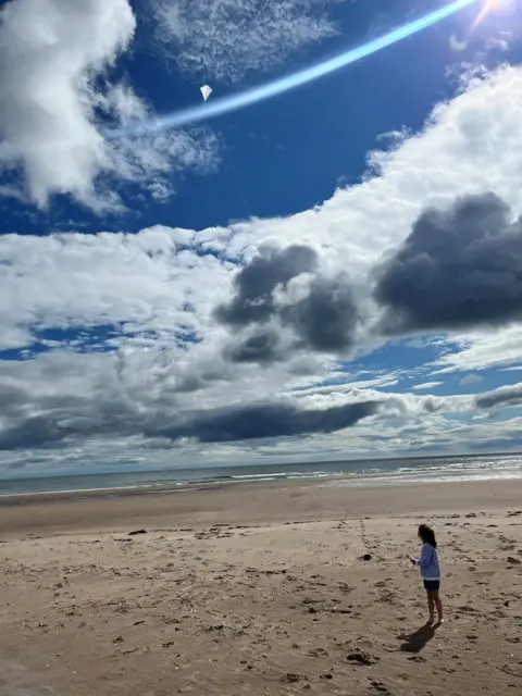 Heather Campbell A young girl standing on a beach, flying a kite into the sky. The clouds are a mixture of white and grey, with the sky bright blue.