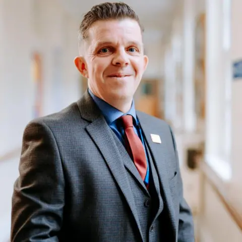 Humber Teaching NHS Foundation Trust A man with short brown hair pushed back and shaved sides is wearing a grey suit with a suit jacket, red tie and blue shirt. He is standing slightly to the side and smiling at the camera. The background is blurred but he is stood in a corridor.