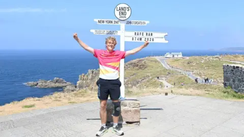 A man stands in front of the Land's End sign, with a sign saying 'Rafe ran 874m 2026' protruding from the side. He has pale skin, blond hair, and is wearing a pink, yellow and white t shirt, with black shorts and a knee support. His arms are outstretched with his hands in fists and he is smiling. Cliffs, the sea, and a white house are visible in the background.
