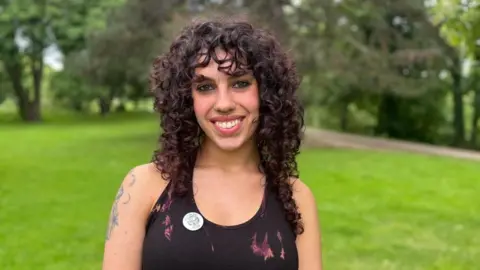 A woman with long black curly hair and a black vest top smiles at the camera. She is standing in a green field with a river path in the background and has a Ross Pride badge attached to her top.