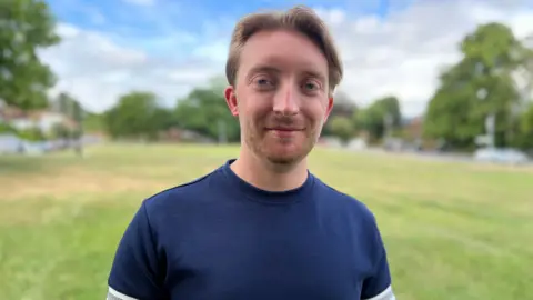 Stuart Woodward/BBC A man standing outside, with a grass common behind him and trees in the background, out of focus. He is wearing a navy blue T-shirt. He is smiling slightly and has strawberry blond hair.