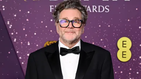Kirk Jones. He has short grey hair and stubble. He is wearing black rimmed glasses, a white shirt, a black suit jacket and a black bowtie. He is looking directly at the camera and smiling.