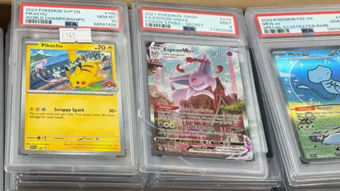 Alex Badham The image shows three damaged Pokémon cards, the clear cases the cards are housed in are cracked and scratched. The card on the left is a yellow Pikachu card with a sticker that says £145 on it, the middle is a pink Espeon card and the card on the left is a blue Mew card, all with cartoon images of the creatures on. 