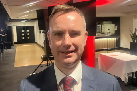 Photograph of Tom Ross, the leader of Trafford Council. The image shows him in a function room at Old Trafford, with chairs and a stage.