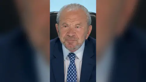 Alan Sugar is wearing a blue suit with a white shirt and a dark blue tie with light blue spots