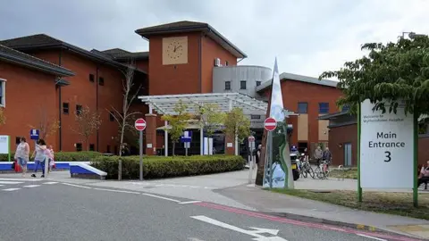 A street view of the main entrance to Wythenshawe Hospital, a redbrick building with a white metal canopy above the public entrance. People can be seen walking out of the site, with a sign bearing the words 'main entrance three' also in view. 