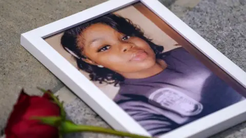 Getty Images A picture of Breonna Taylor, who is wearing a purple top and has black hair, in a white picture frame with a red rose lying next to it
