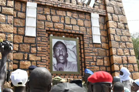 AFP Pessoas olhando para uma placa com um retrato do revolucionário de Burkina Faso, Thomas Sankara, em uma parede de tijolos