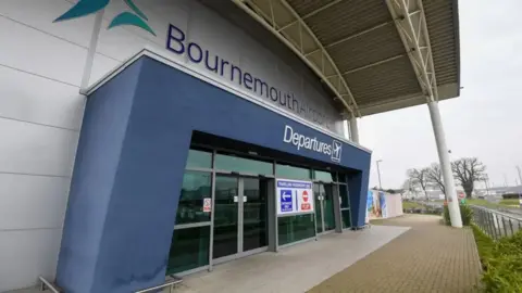 Exterior view of the entrance to Bournemouth Airport departures  - a grey building with glass doors set in a blue arch with the words 'Bournemouth Airport' above