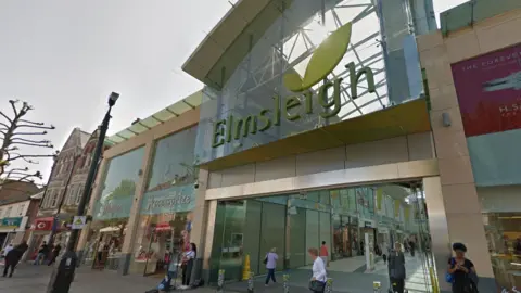 Google Outside of a shopping centre. There are people walking in and out of the entrance. Above the entrance is lots of glass, on which there is the word Elmsleigh written in green lettering