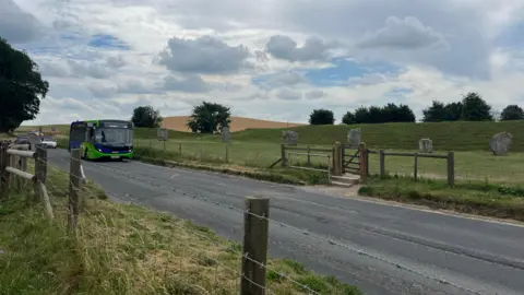 The road running through Avebury with traffic, including a bus, approaching. Behind the road can be seen a field with part of the large stone circle visible, and behind those stones is a grassy embankment with trees in the distance. 