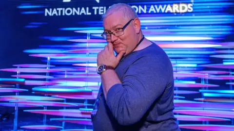 Rick Bronks James Kenny is standing sideways in front of a blue and purple promotional screen at the National Television Awards, with the NTA branding behind him. He is a white man with short grey, thinning hair who is smiling and holding his hand up to his chin in a comedic way. He is wearing a blue jumper, square glasses and a NTA lanyard.