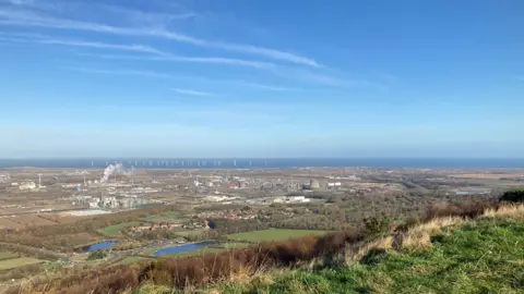 The Wilton International industrial complex is a collection of industrial buildings surrounded by fields. Smoke is rising form one of them. The sea can be seen in the background, with wind turbines off the coast.