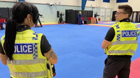 Two police officers wearing uniform and high visibility police vests standing in the training hall at Wiltshire Police's headquarters in Devizes