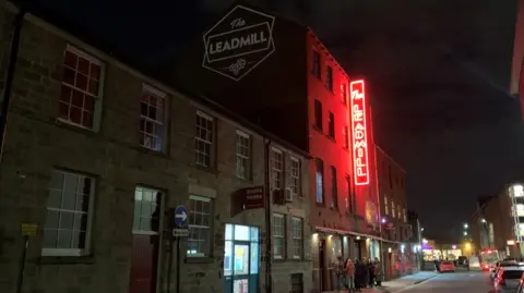 A four-storey red brick building with a black sign running vertically along the wall with the words 'The Leadmill' in red, neon writing. 
