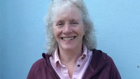 MANX DEAF SOCIETY Pam Ashcroft, a woman with curly grey hair and a fringe smiles, she wears a pink shirt and stands in front of a blue wall.