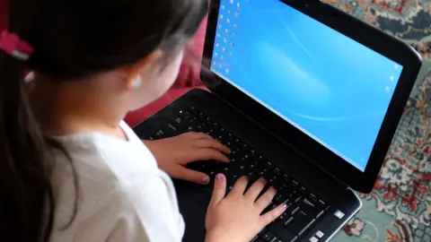 PA Media A child with long dark hair and sat on a colourful, patterned carpet, uses a laptop computer, currently on a desktop screen.