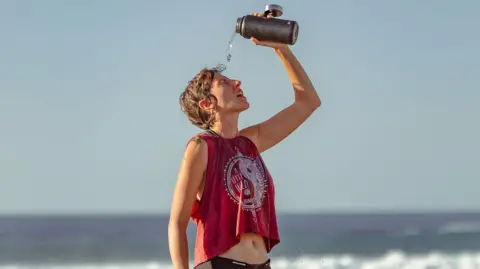 Getty Images A person is standing on a shingle beach pouring water from a flask onto their head. They are wearing black shorts and a cropped dark red sleeveless top with a white logo. The sky is cloudless and waves can be seen breaking in the background.