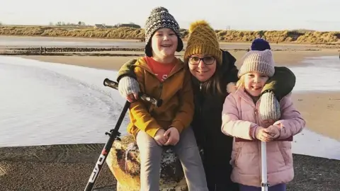 Esther Graham Esther Graham is in her thirties and is on a pier, crouching down with her arms around her two children who are about three or four years old. She is wearing a yellow hat, has glasses and a brown jacket. The boy, with a yellow jacket and grey bobble hat, is sitting on a concrete bollard. The girl, who is wearing a pink jacket and hat is standing beside it. Behind the trio there is a big beach with the sand dunes fringing it.