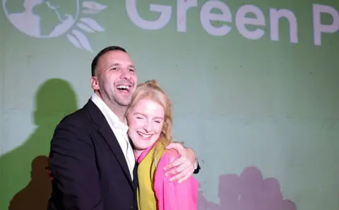 Getty Images Grinning Green Party leader Zack Polanski hugs victorious candidate Hannah Spencer against a Green Party backdrop
