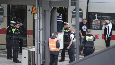 Police officers standing on a train platform, in front of a train, and a couple of members of the public standing nearby