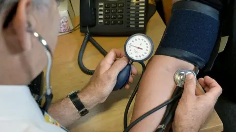 A doctor measuring the blood pressure and the heart rate of a patient. The patient's sleeve is rolled up and there is a pressure pad around the arm. The doctor is holding a stethoscope on the arm and reading a gauge.