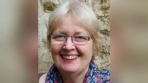 Family A woman with short blonde hair, glasses, and a blue and purple scarf, smiles at the camera.