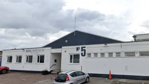 Google A large single-storey white building with a grey roof and the words "Soul Survivor Watford" and "No 5" painted in grey on the front. There are two cars parked outside.