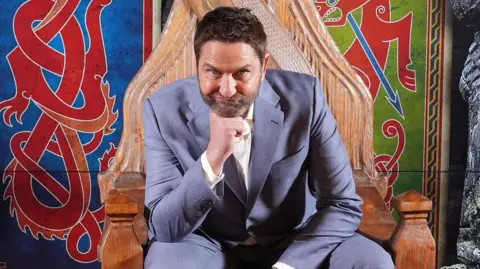 A man in a light grey-blue suit and a white shirt is sitting on a wooden throne, in front of a Viking-style mural. He is resting his chin on his fist.