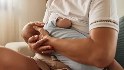 A woman wearing a white t-shirt cradles a baby wearing a light blue outfit as they breastfeed. The woman is sitting on a light coloured chair or sofa with a light coloured curtain in the background.
