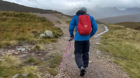 Robert Marshall The picture is taken behind of Robert walking along the West Highland Way. Its a bleak and dramatic sky with grass on either side of the gravel path. He is wearing a blue jacket, red rucksack and black trousers. He is carrying purple walking poles.