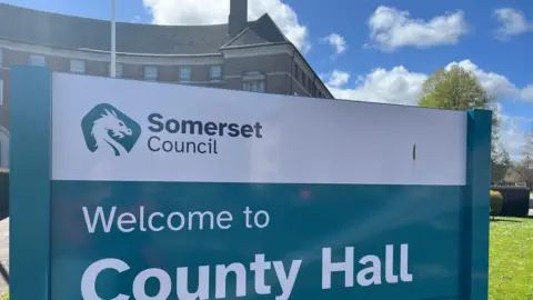 BBC The sign for Somerset Council's county hall. It is blue and white. In the background is part of the building and some grass.