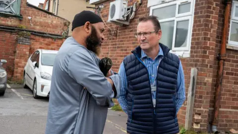 OFPCC A man in a blue robe and black head covering speaks to a man with glasses, a blue and white striped shirt and a navy gilet in a car park.