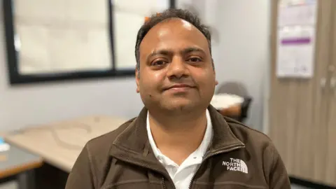 JOANNA TAYLOR/BBC Abhishek Srivasdava smiling and looking at the camera. He is wearing a white shirt and brown fleece. There is an out of focus classroom behind him. 