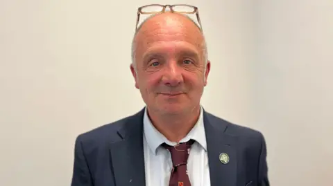 Deputy Neil Inder - A bald man with glasses on his head. He is smirking at the camera, and has wrinkles around his blue eyes. He is wearing a blue jacket and a white shirt with a purple tie. 