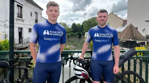 James Howell on the left with riding partner Max Edginton. They are each wearing blue and white cycling clothes and are stood in front of their bikes