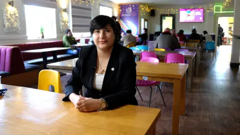 Kate Bradbrook/BBC Zahra Karimi sits at a canteen table with her hands folded and smiles at the camera. She has short black hair and wears a dark smart jacket over a white top, and wears a gold wrist watch.