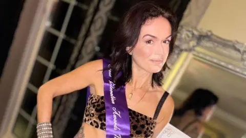 A woman with shoulder-length brown hair looks at the camera while wearing a cropped leopard print top. She also wears a purple sash with white writing on it.