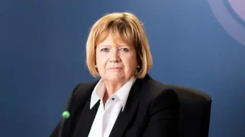 Pirahna Photography Baroness Hallett in front of a blue backdrop featuring the pink, purple and white Covid-19 inquiry logo. 